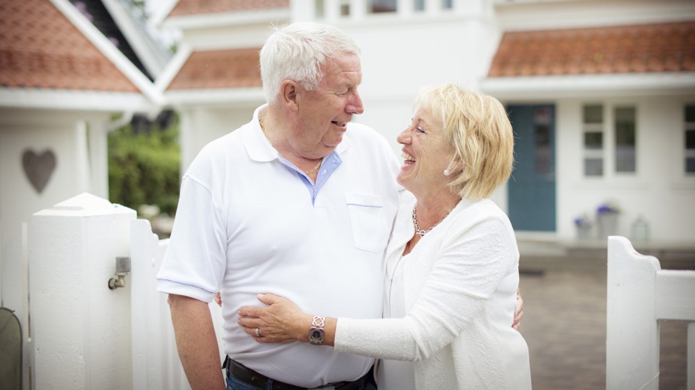 En gammel mann og kvinne holder armene rundt hverandre mens de ser på hverandre og smiler